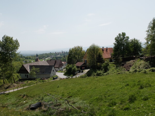 Blick von einem Hang im Vordergrund auf eine ländliche Siedlung mit Bauernhöfen und Häusern, umgeben von Wiesen, Bäumen und sanften Hügeln im Hintergrund.
